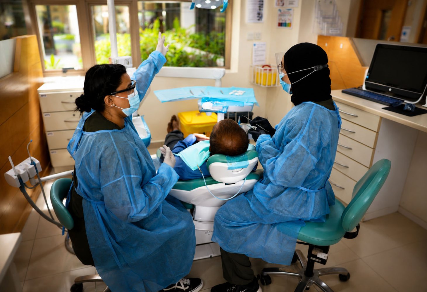 Two RAKMHSU dental students in blue gowns and face masks working either side of a patient reclined in the chair at the college dental clinic