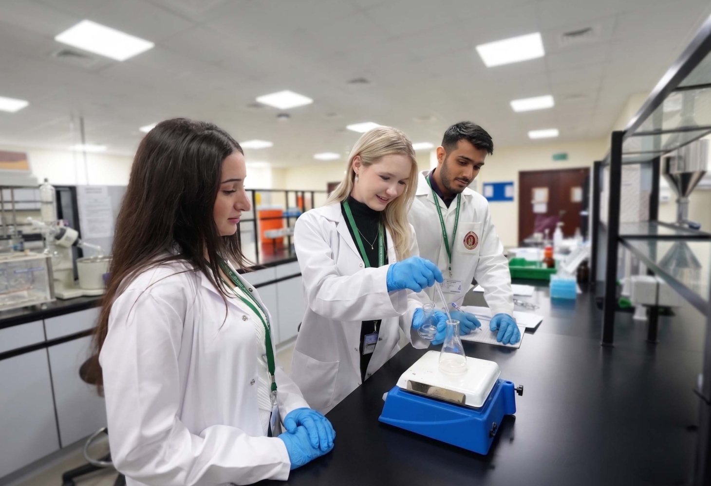 Three RAKMHSU pharmacy students in lab coats and gloves working with a conical flask on a hotplate stirrer in the pharmaceutics laboratory
