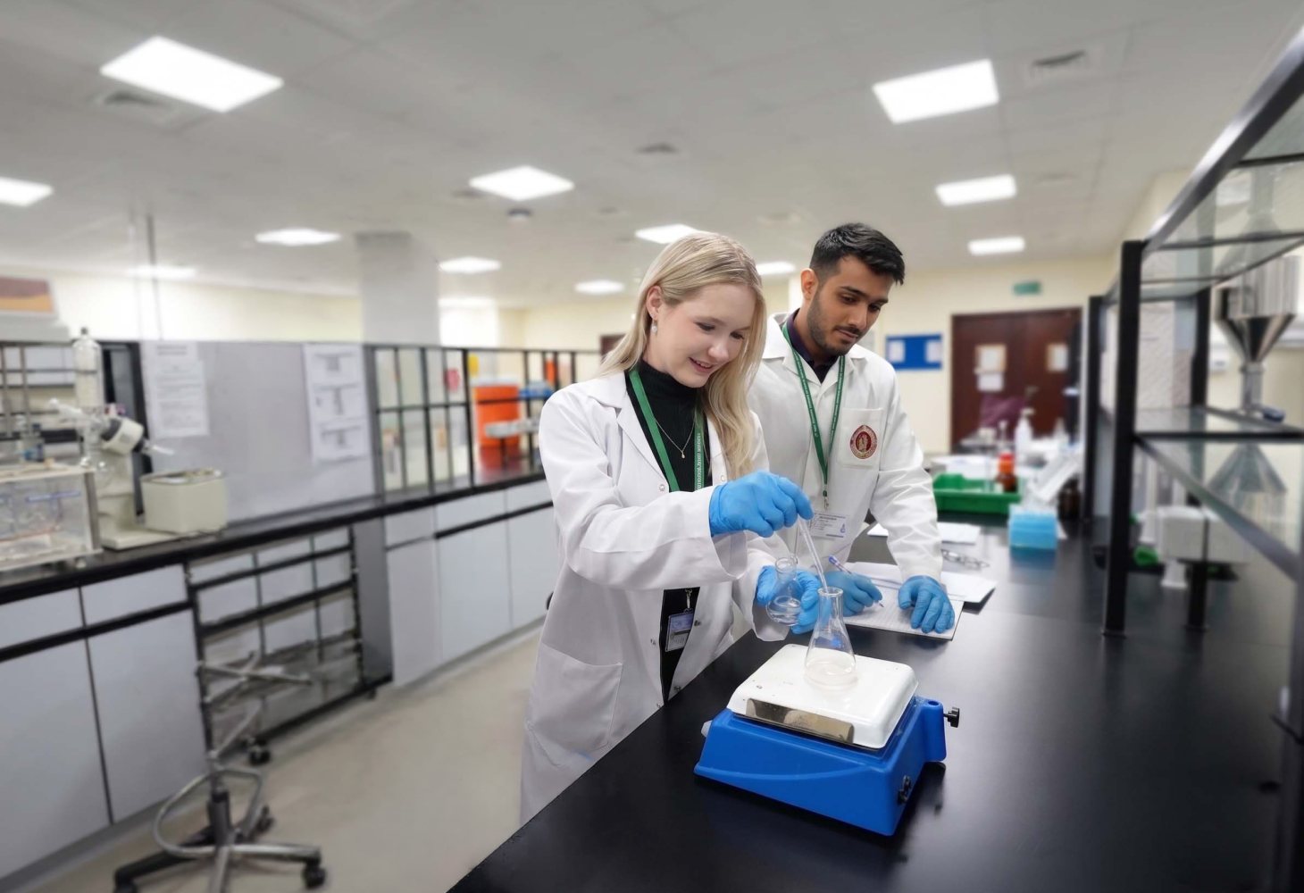 Two RAKMHSU pharmacy students in lab coats and blue gloves preparing a solution in a conical flask on a hotplate stirrer in the pharmacy laboratory