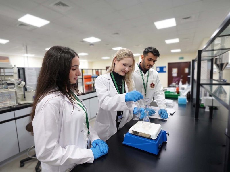 Three RAKMHSU pharmacy students in lab coats and gloves working with a conical flask on a hotplate stirrer in the pharmaceutics laboratory