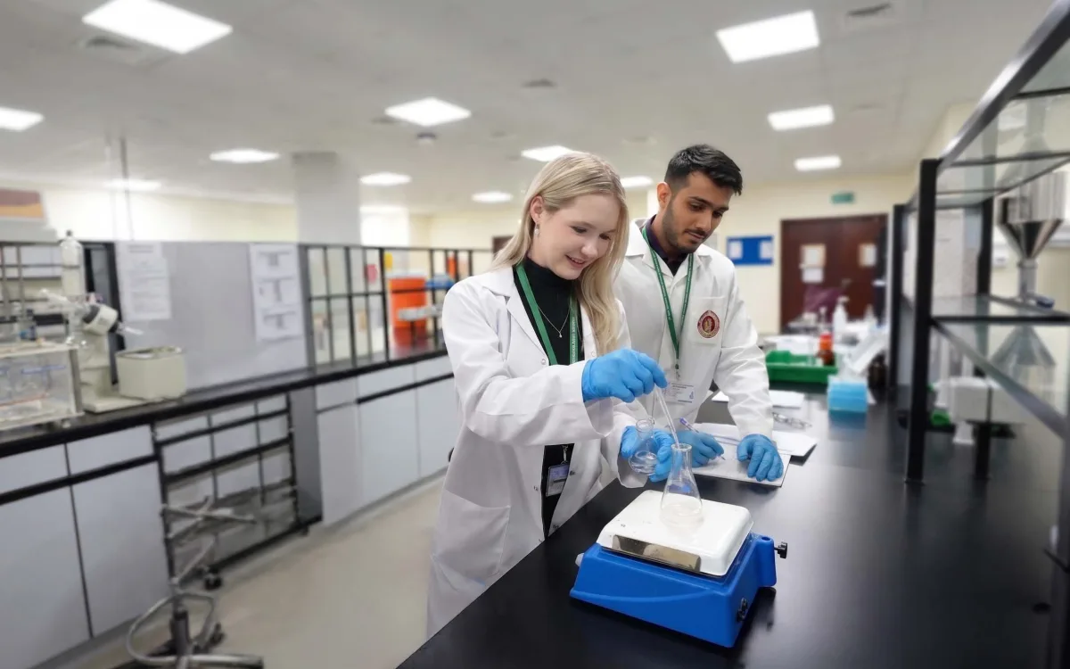 Two RAKMHSU pharmacy students in lab coats and blue gloves preparing a solution in a conical flask on a hotplate stirrer in the pharmacy laboratory