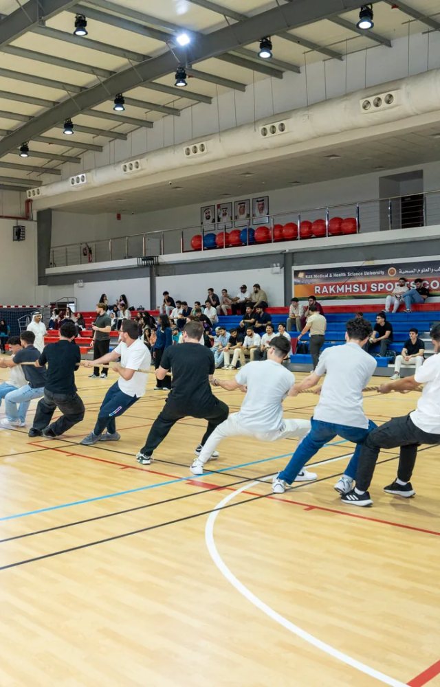 Students hauling on the rope in a tug-of-war on the court at the RAKMHSU Sports Complex, watched by spectators in the bleachers