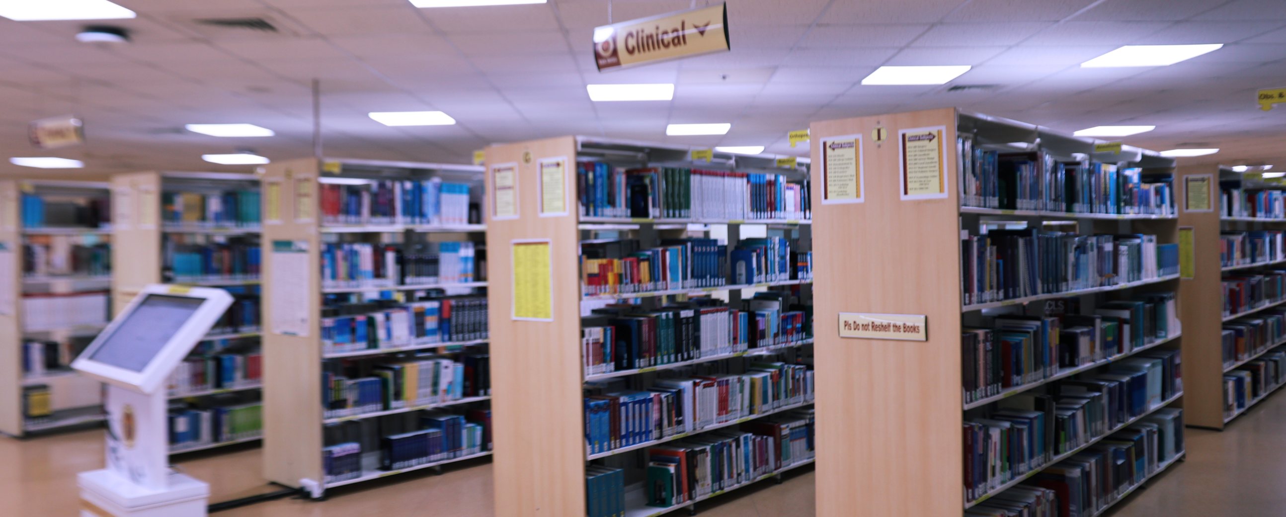 Book stacks in the RAK MHSU Library, with the Clinical collection signposted overhead and a catalogue search kiosk at the end of the aisle