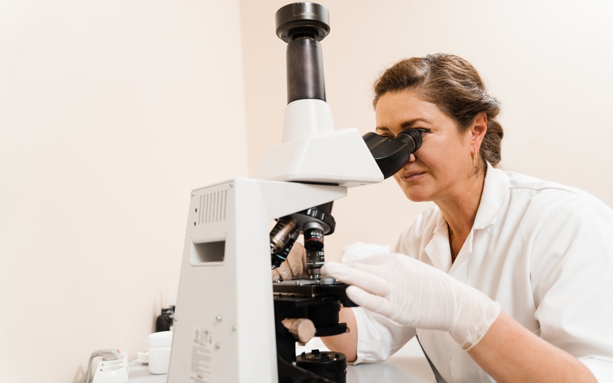 A researcher in a white coat and gloves looking through a laboratory microscope