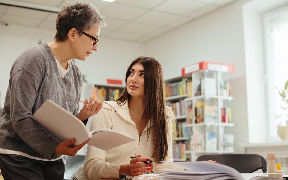 A senior academic holding an open book and talking with a student seated at a library desk
