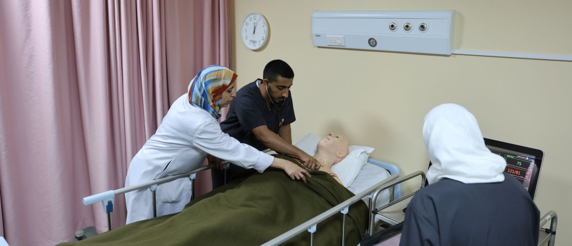 A nursing instructor and two students at a simulation bedside, one student listening to the chest of a high-fidelity patient manikin with a stethoscope while the vital-signs monitor beside the bed shows a heart rate and blood pressure