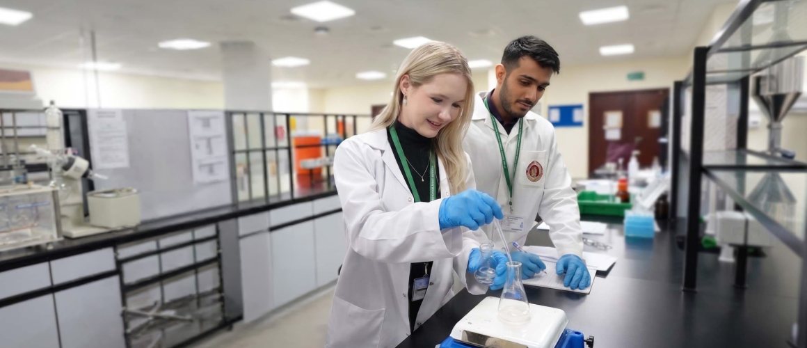 Two RAKMHSU pharmacy students in lab coats and blue gloves preparing a solution in a conical flask on a hotplate stirrer in the pharmacy laboratory