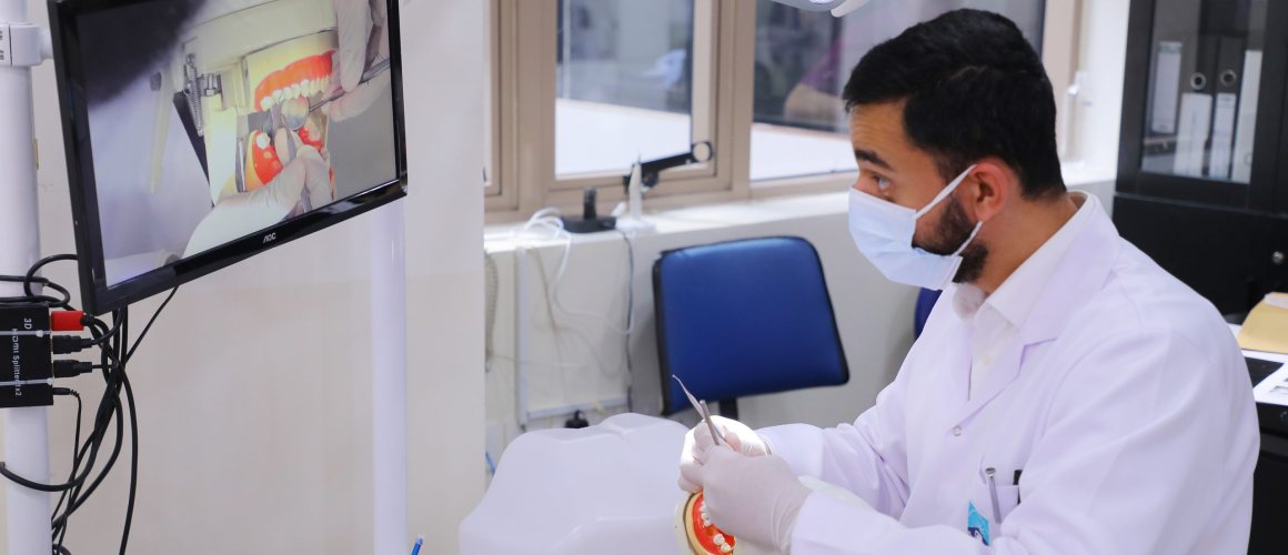 A masked clinician in a white coat working on a dental phantom head with a hand instrument in the RAKMHSU simulation clinic, the intraoral view relayed on a monitor beside the chair