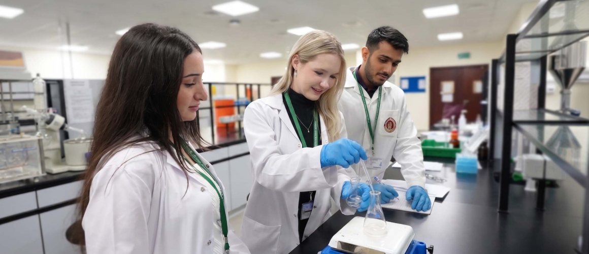 Three RAKMHSU pharmacy students in white coats and gloves working at a laboratory bench, one pouring a solution into a conical flask on a magnetic stirrer while another records the result