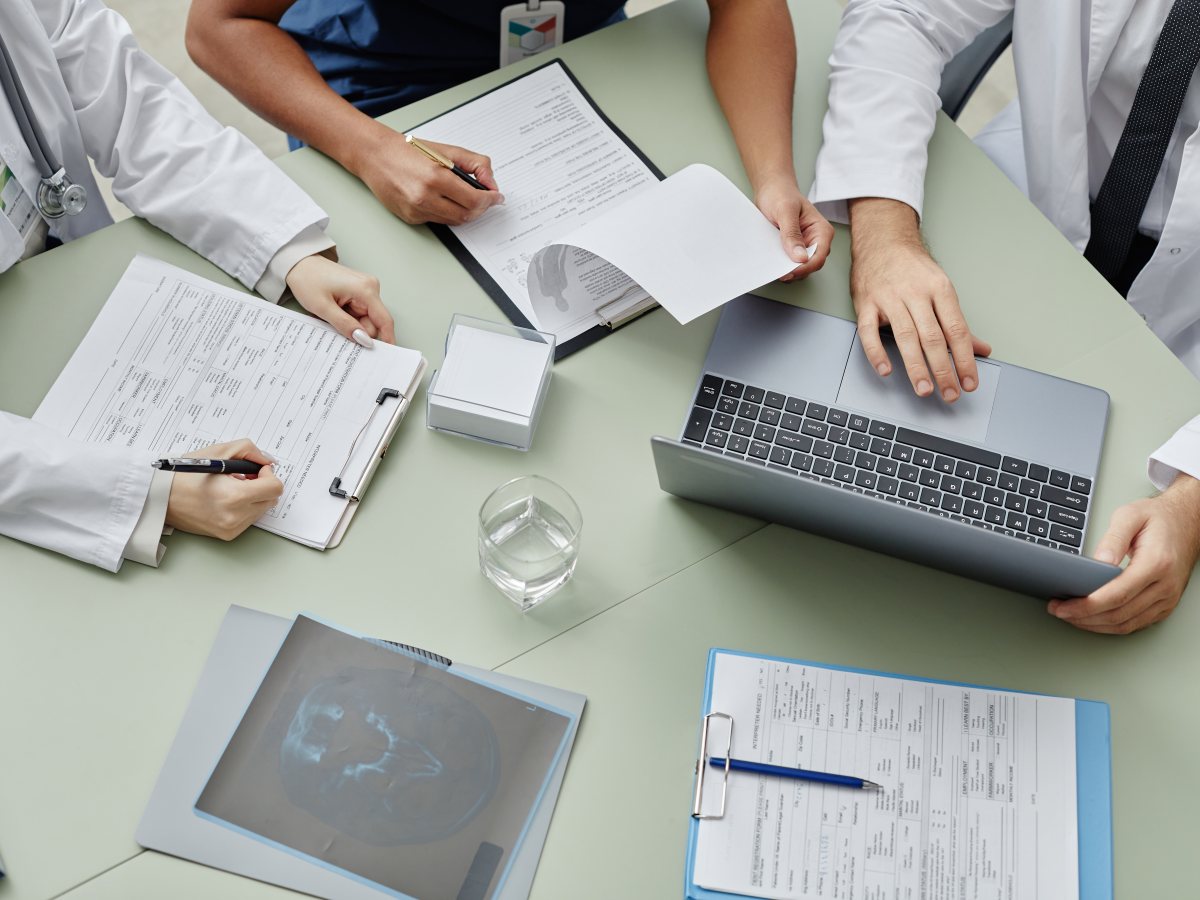 Clinicians at a meeting table reviewing patient charts, an X-ray and a laptop, seen from above