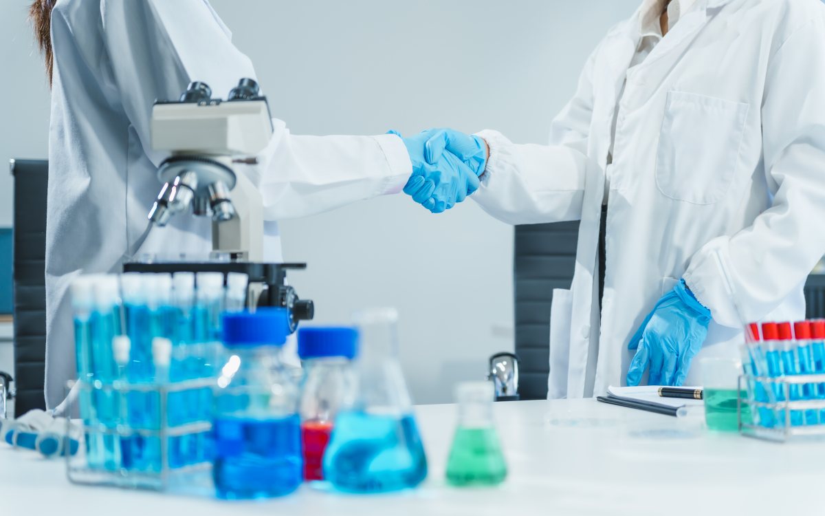 Two researchers in laboratory coats and blue gloves shaking hands over a bench of glassware