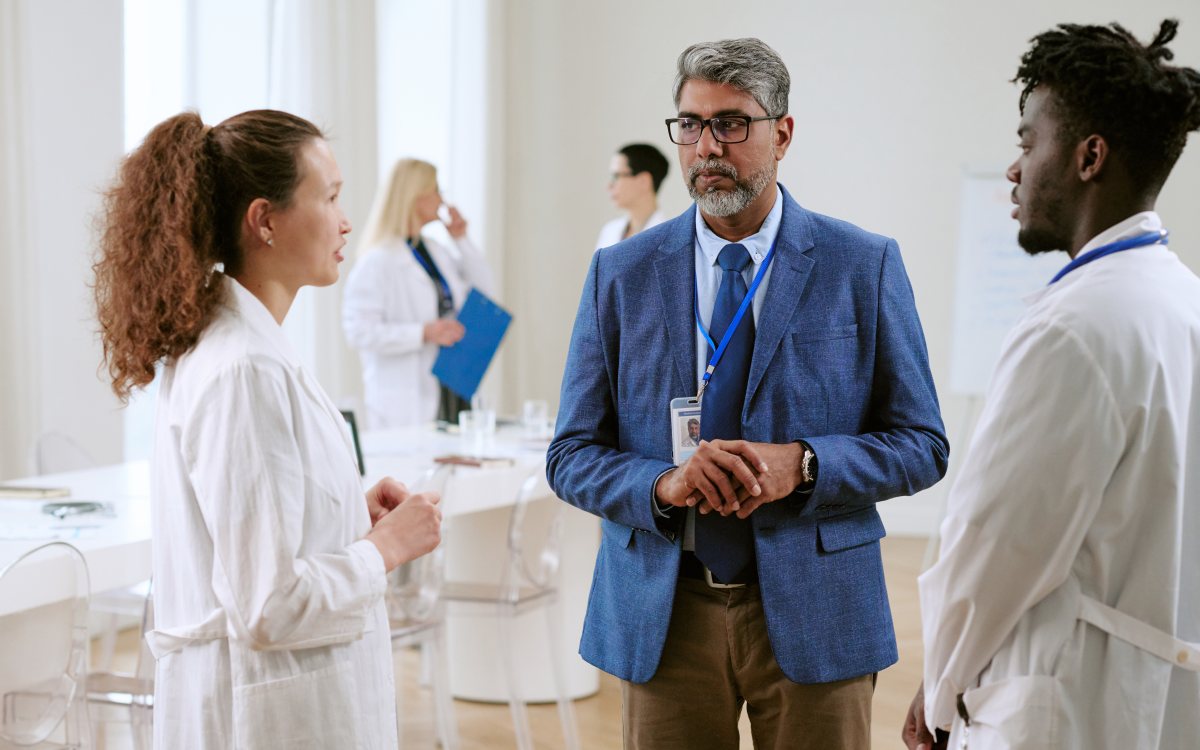 A man in a blue blazer talks with two young researchers in white laboratory coats, with colleagues behind them