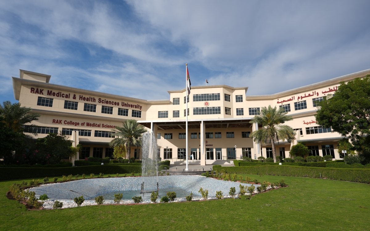 The main building of RAK Medical & Health Sciences University under a blue sky, with the UAE flag at its entrance and a fountain on the front lawn